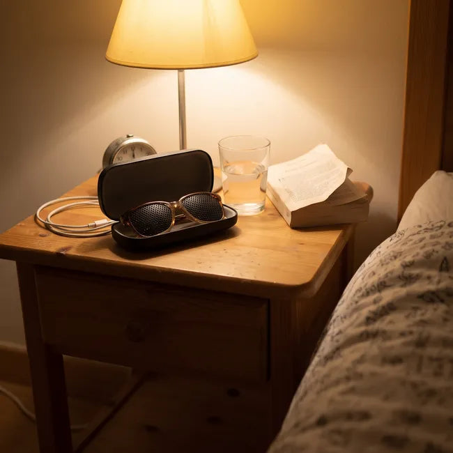 Table de chevet le soir avec lampe allumée, réveil et étui ouvert contenant des lunettes à trous